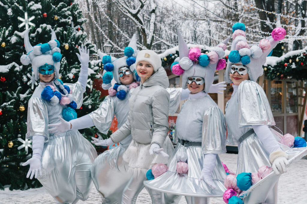 Group of performers in festive silver outfits with colorful pom-pom headgear, posing in a snowy winter setting with Christmas trees in the background.
