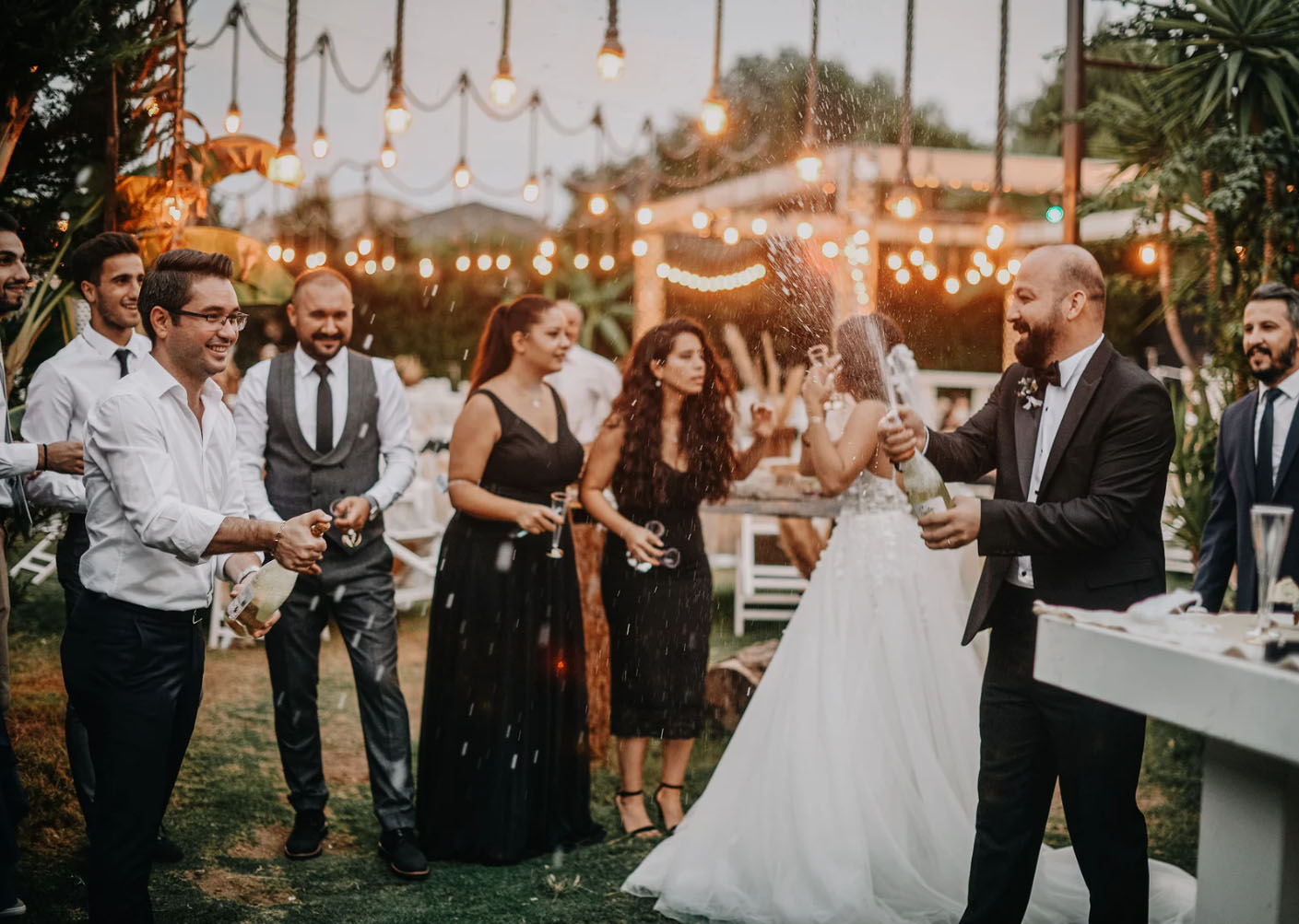 A group of people celebrating at a wedding, with a man in a tuxedo spraying champagne while others smile and watch in the beautifully lit outdoor setting.