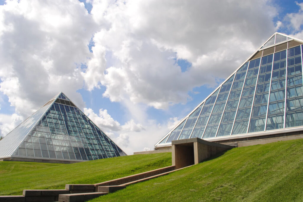 Two large glass pyramids at the Muttart Conservatory in Edmonton, surrounded by green grass under a bright blue sky with scattered clouds.