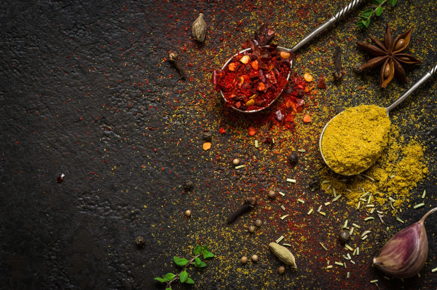 A close-up view of various spices scattered on a dark surface, including red chili flakes, turmeric powder, star anise, and cardamom, with two metal spoons.