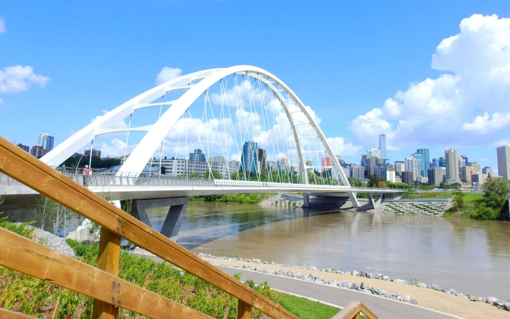 The Walterdale Bridge in Edmonton, with its modern white arch design, crossing the North Saskatchewan River, with the city's skyline visible in the background and a clear blue sky.
