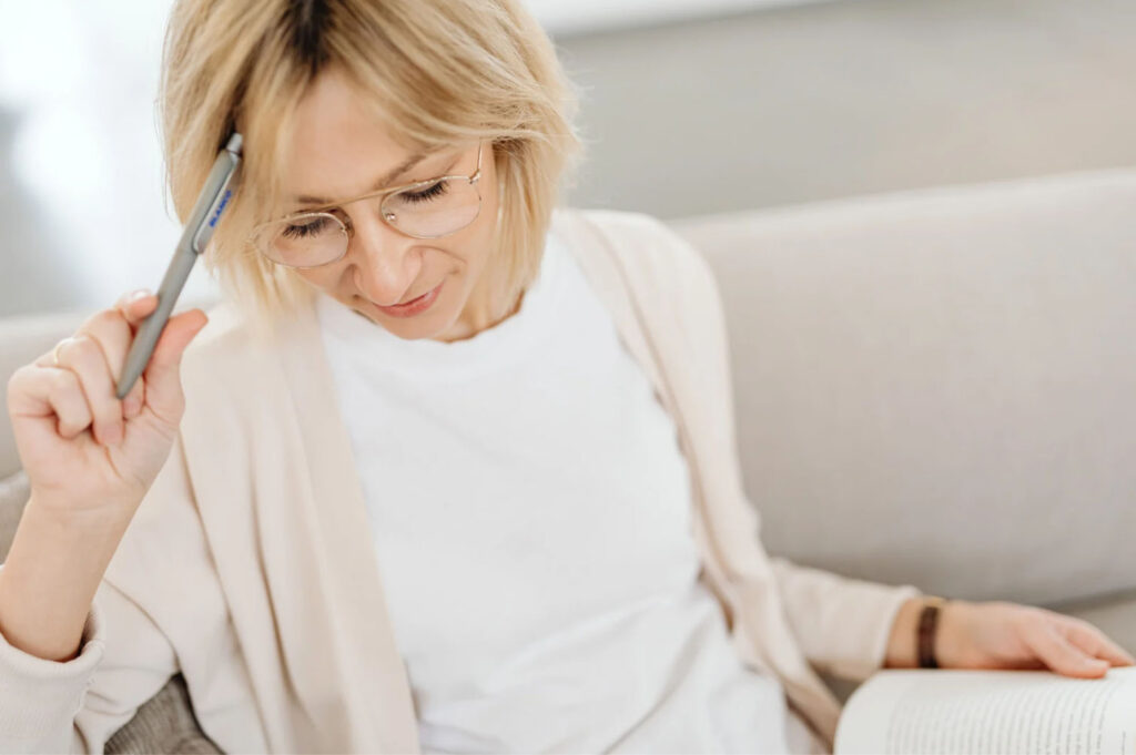 A woman sitting on a couch with a book in her lap, holding a pen and resting it against her head, appearing thoughtful or deep in concentration.