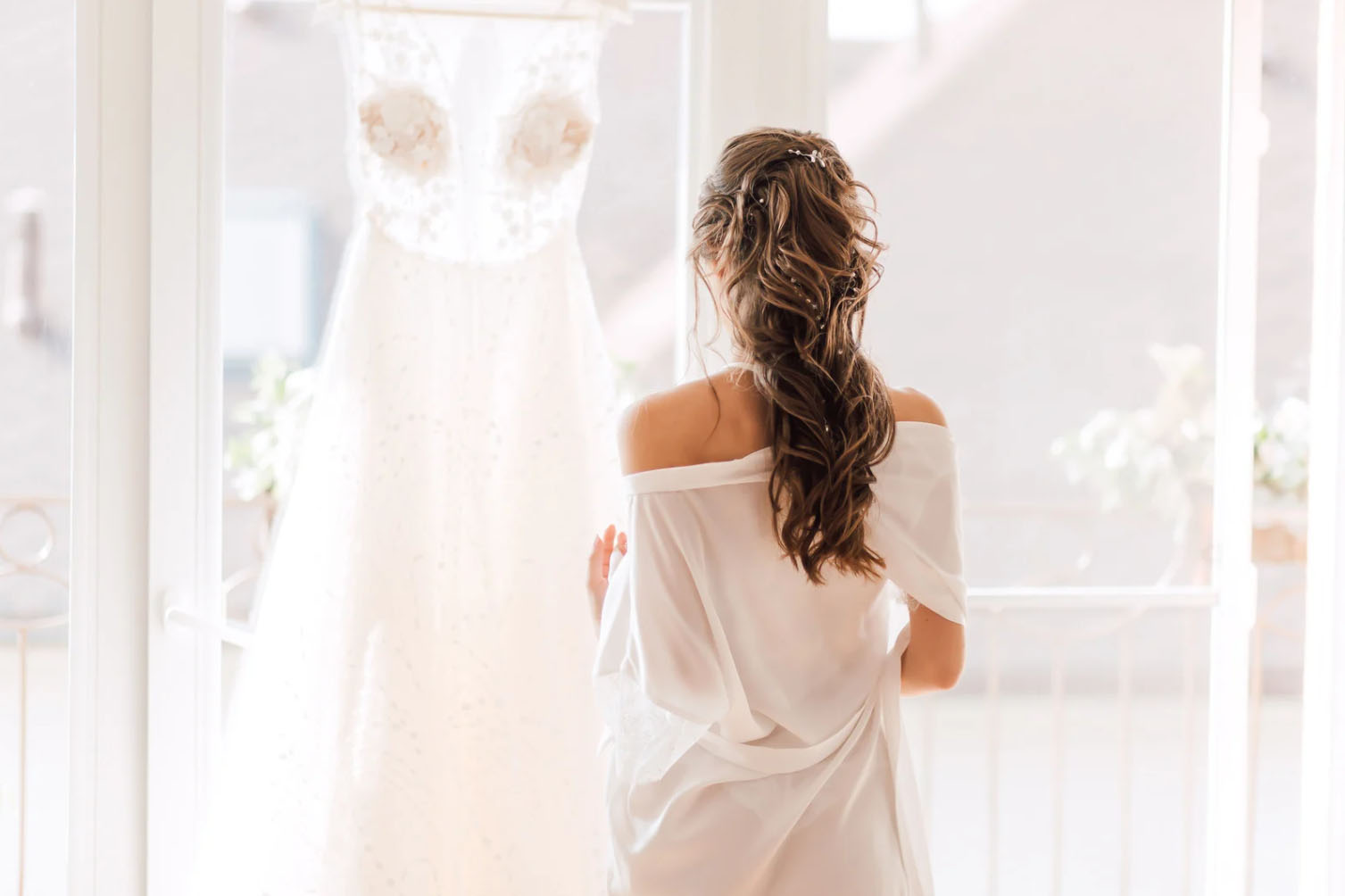 A woman in a white robe stands in front of a window, gazing at a wedding dress hanging on a hanger. The scene is serene and captures a moment of anticipation before the wedding.