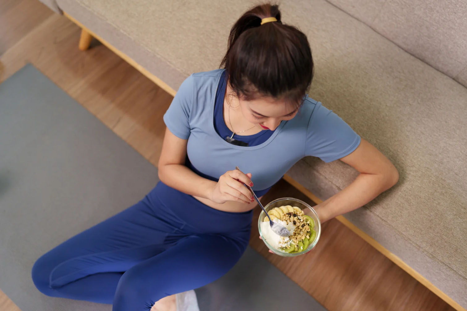 A woman in athletic wear sitting on a yoga mat, enjoying a healthy bowl of yogurt topped with fruit and granola. She is focused on her meal after a workout.
