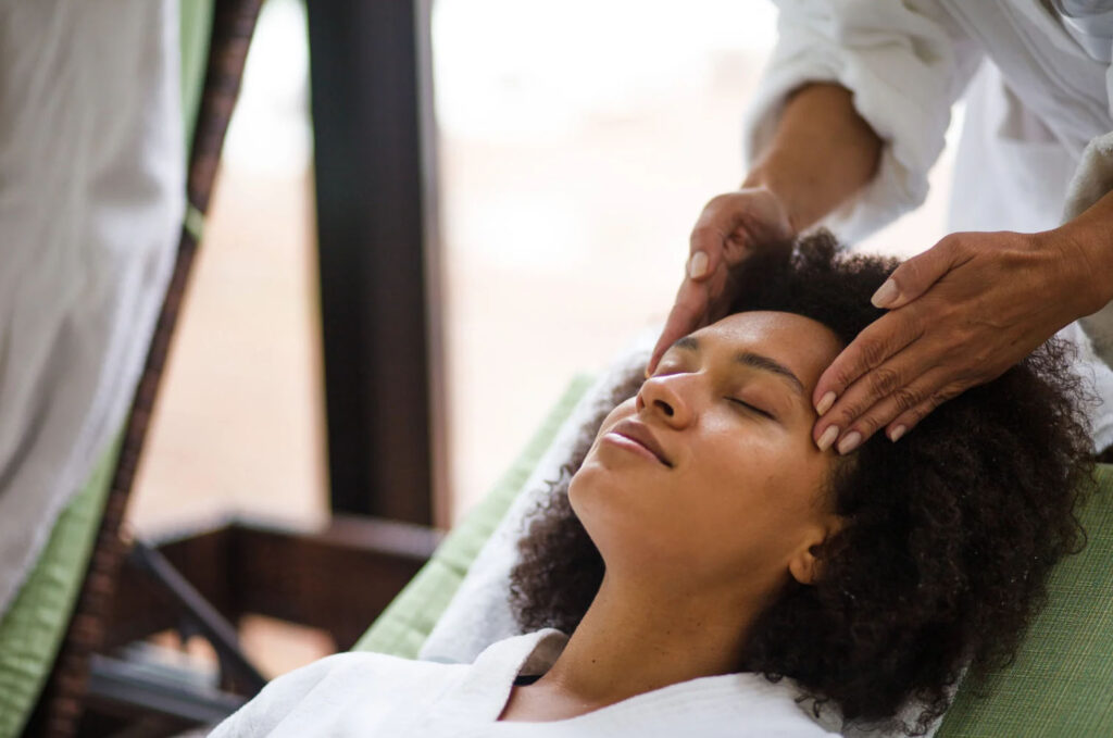 A woman with curly hair relaxing during a spa treatment, receiving a gentle head massage from a therapist in a serene setting.