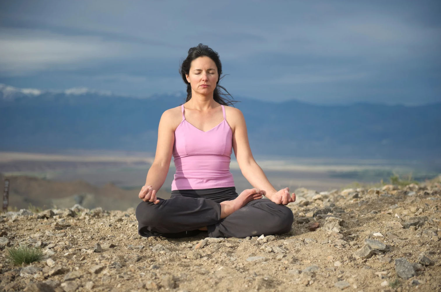 A woman practicing meditation in a lotus position outdoors on a rocky surface, with a serene mountain landscape in the background.