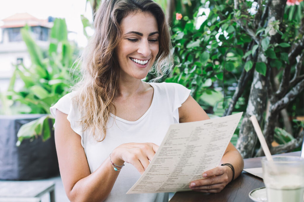 A woman smiling and looking at a menu while sitting in a café with greenery in the background. She is holding the menu in her hands, wearing a white shirt.