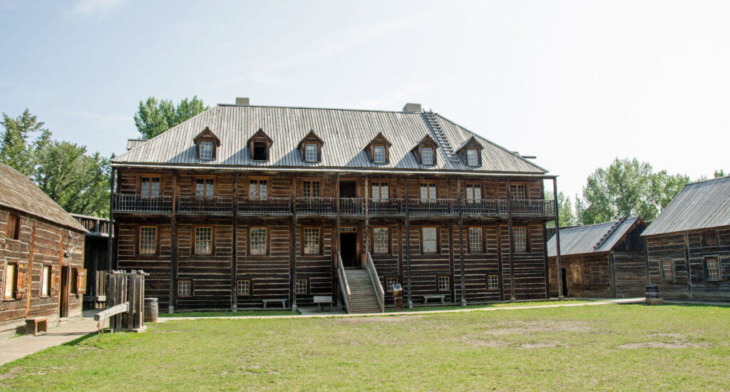 A historic wooden building with multiple stories, situated in a grassy area surrounded by smaller wooden structures, representing the pioneer era at Fort Edmonton Park.