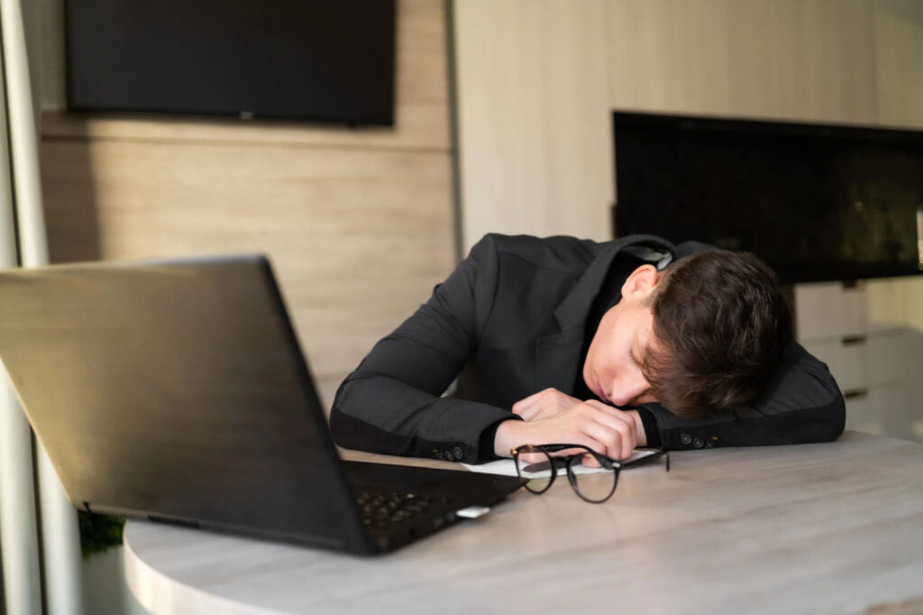 A young man in a suit resting his head on his desk next to a laptop, showing signs of exhaustion or fatigue.