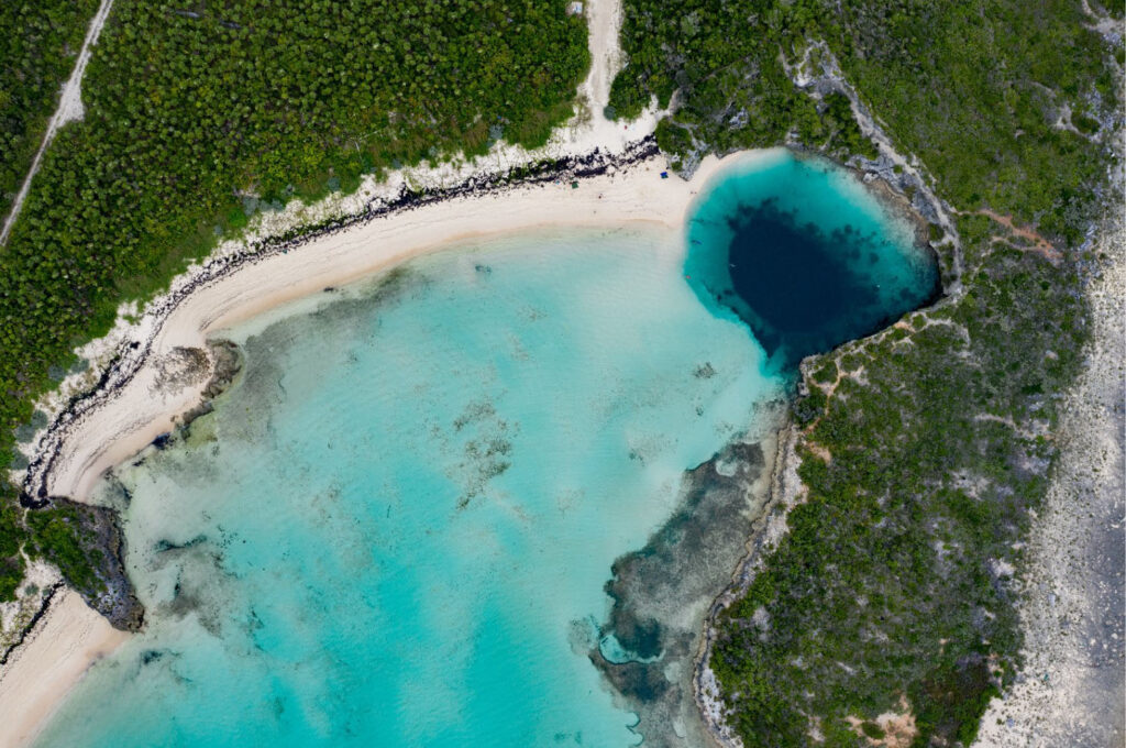 Aerial view of Dean’s Blue Hole on Long Island, Bahamas, showing a deep circular blue sinkhole beside a white sand beach and shallow turquoise waters surrounded by lush green vegetation.