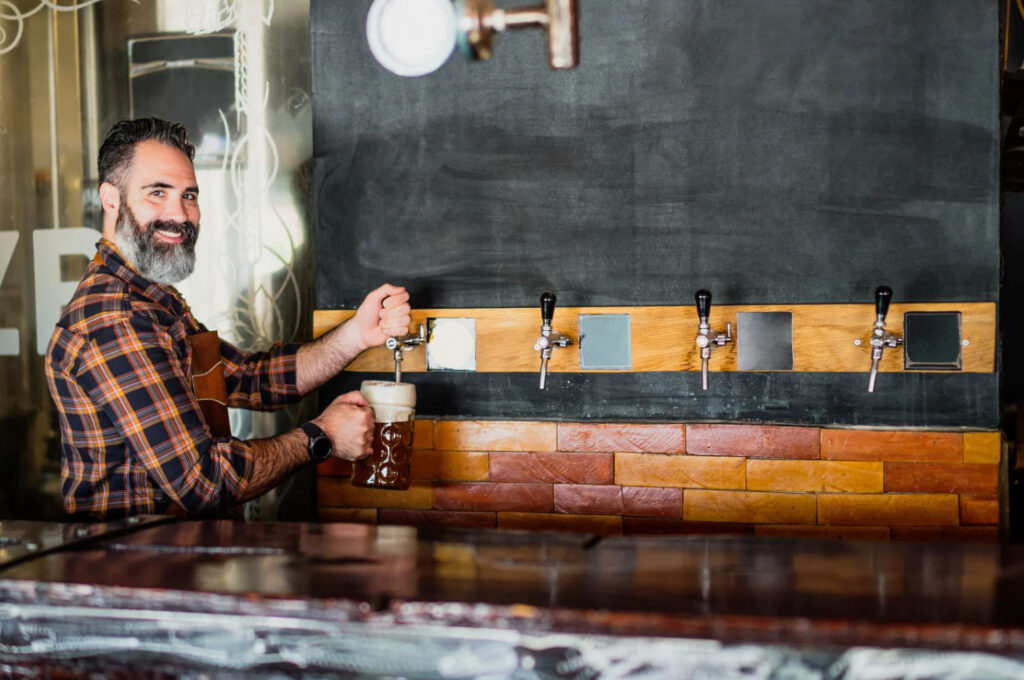 A bearded man in a plaid shirt pouring beer from a tap into a glass mug at a bar with multiple beer taps on the wall behind him.