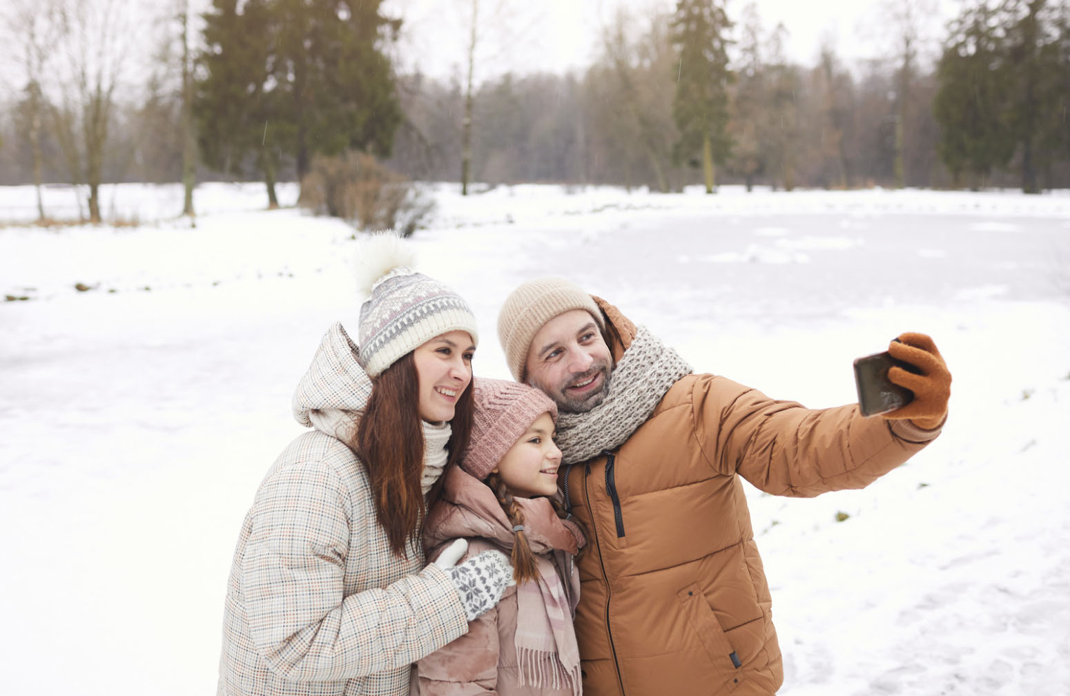 A family of three, dressed warmly in winter coats and hats, is smiling and taking a selfie in a snowy outdoor setting. The scene captures a joyful winter moment together.