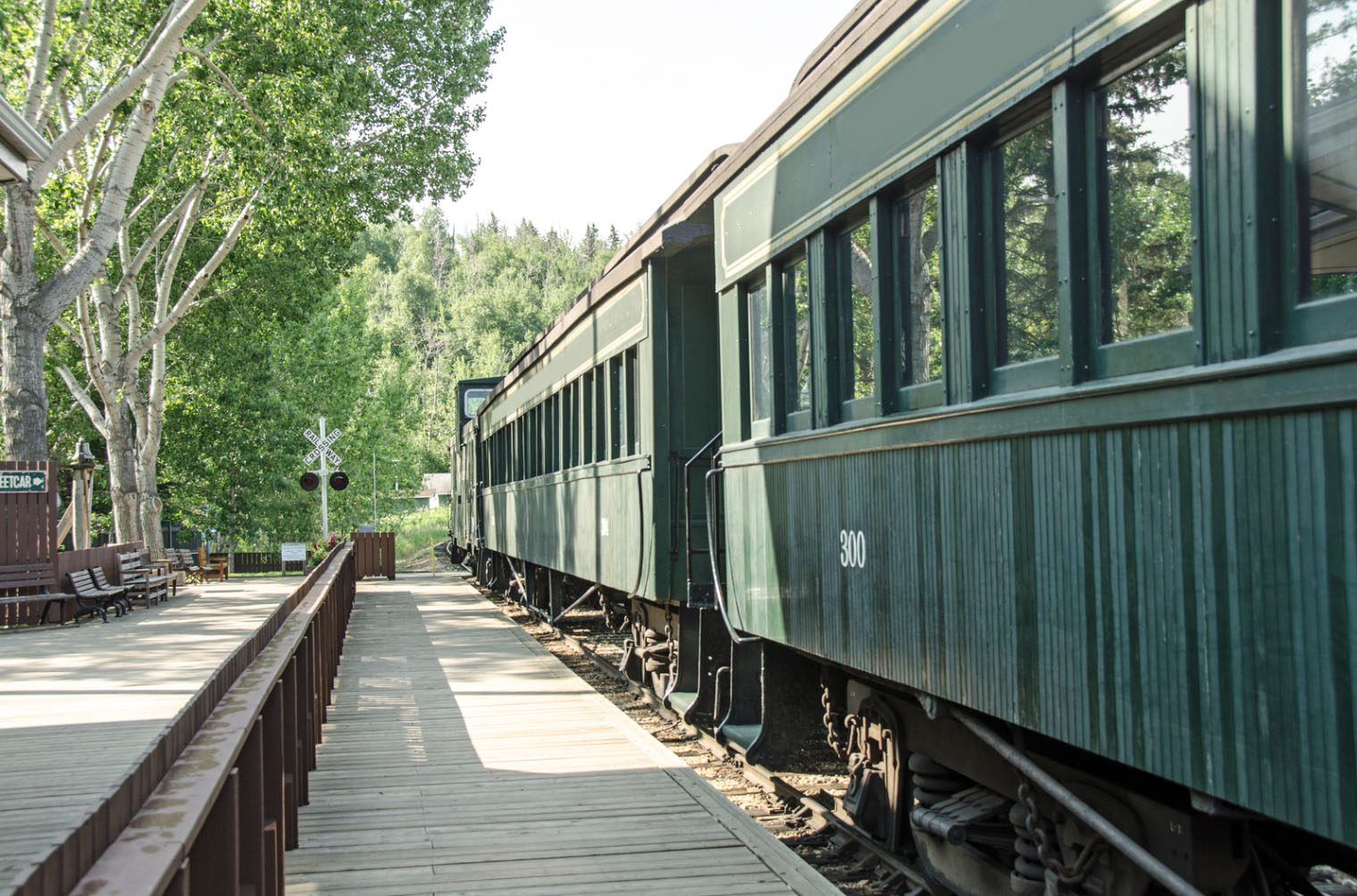A historic green train car labeled "300" parked at a wooden platform surrounded by trees, with a railroad crossing signal in the background.