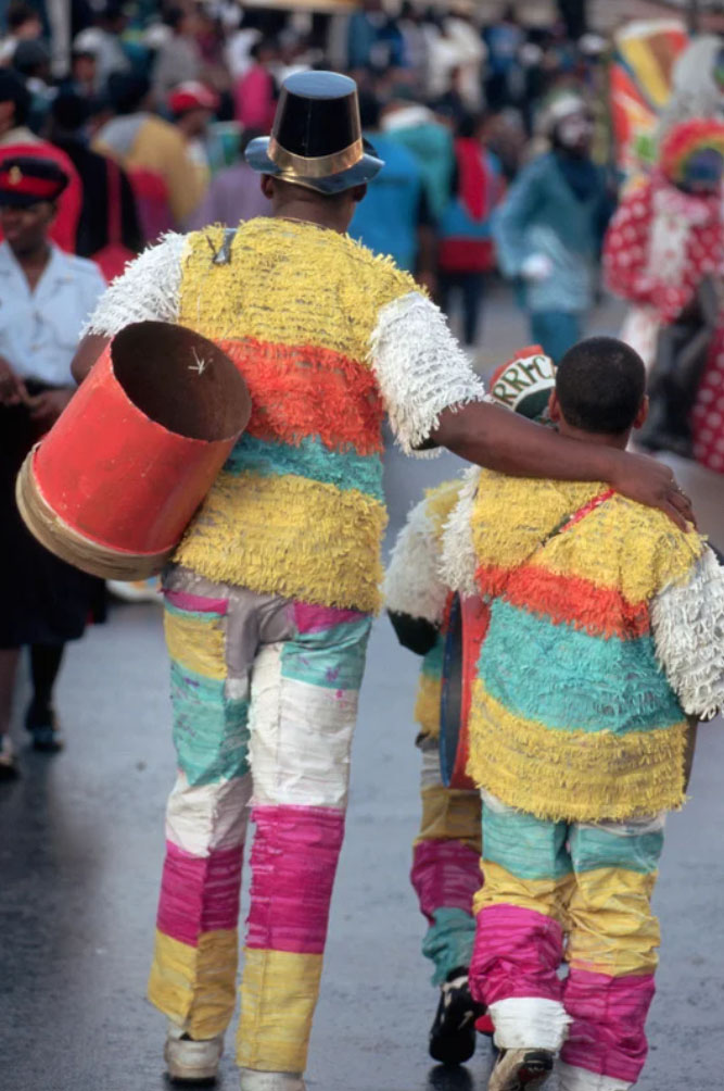 Two participants in colorful Junkanoo costumes walk arm in arm through a Nassau street parade, wearing fringed outfits and carrying a drum amid a lively festival crowd.