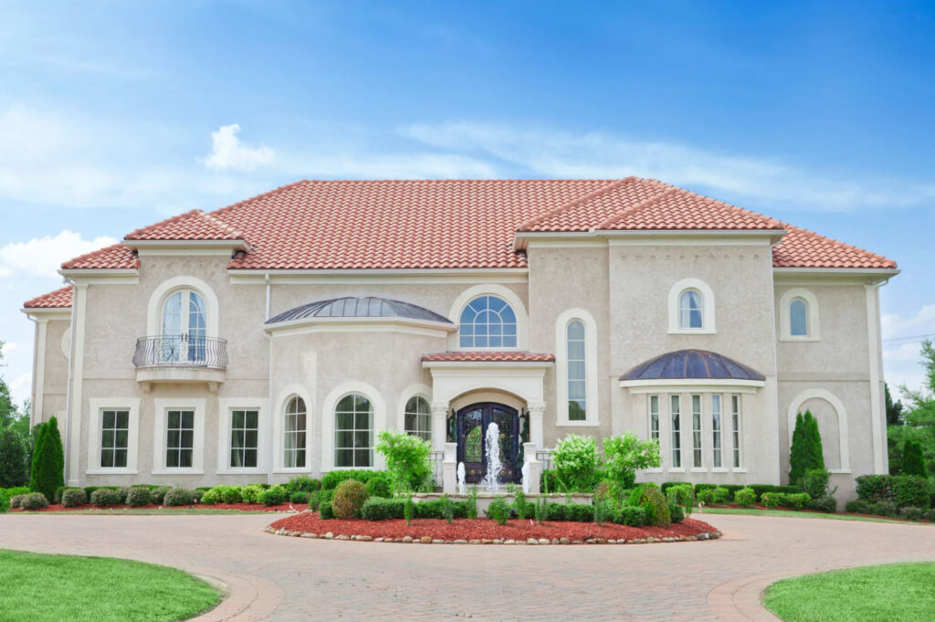 A large luxury Mediterranean-style mansion with a red tile roof, arched windows, and a landscaped circular driveway under a clear blue sky.