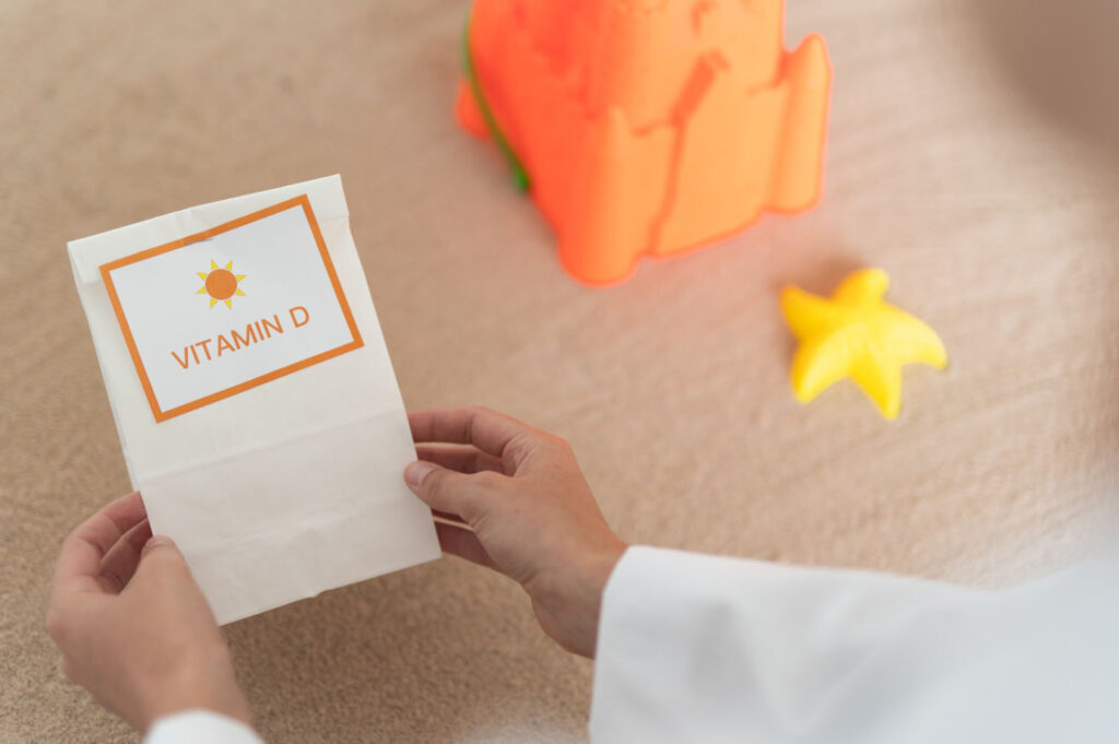 A person holds a paper bag labeled "Vitamin D" with a sun symbol, placed on a light surface with a toy sandcastle and a starfish nearby, suggesting a sunny, beachy theme.