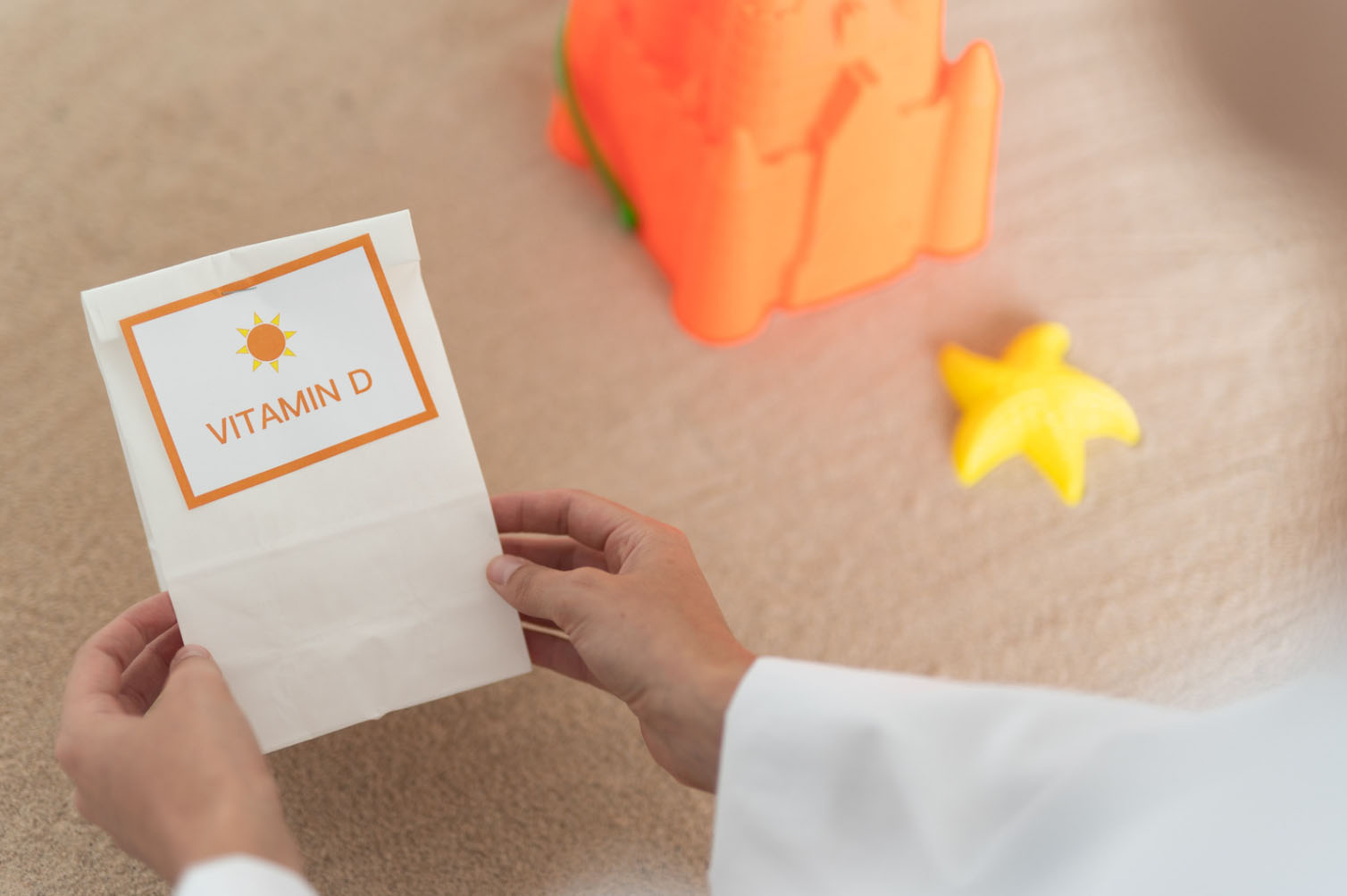 A person holds a paper bag labeled "Vitamin D" with a sun symbol, placed on a light surface with a toy sandcastle and a starfish nearby, suggesting a sunny, beachy theme.