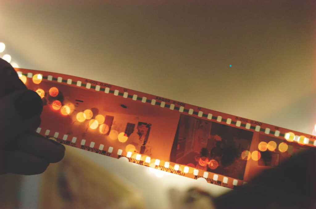 A person holding a strip of developed photographic film against warm light, with visible film frames and soft bokeh highlights in the background.