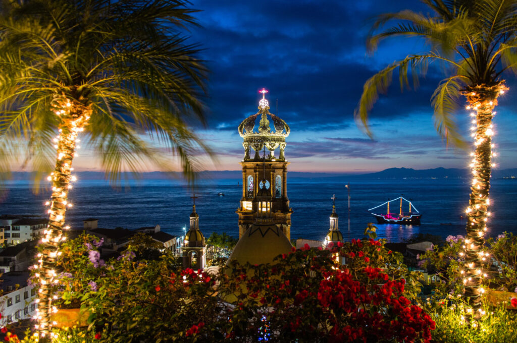 Nighttime view of Puerto Vallarta’s illuminated church tower framed by palm trees, overlooking the ocean with a lit boat on the water at dusk.