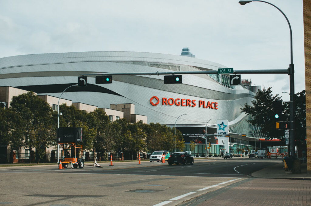 A view of Rogers Place, a modern arena in Edmonton, Canada, with a clear sky, street traffic, and green trees in the foreground. The large Rogers Place sign is prominently displayed on the building.