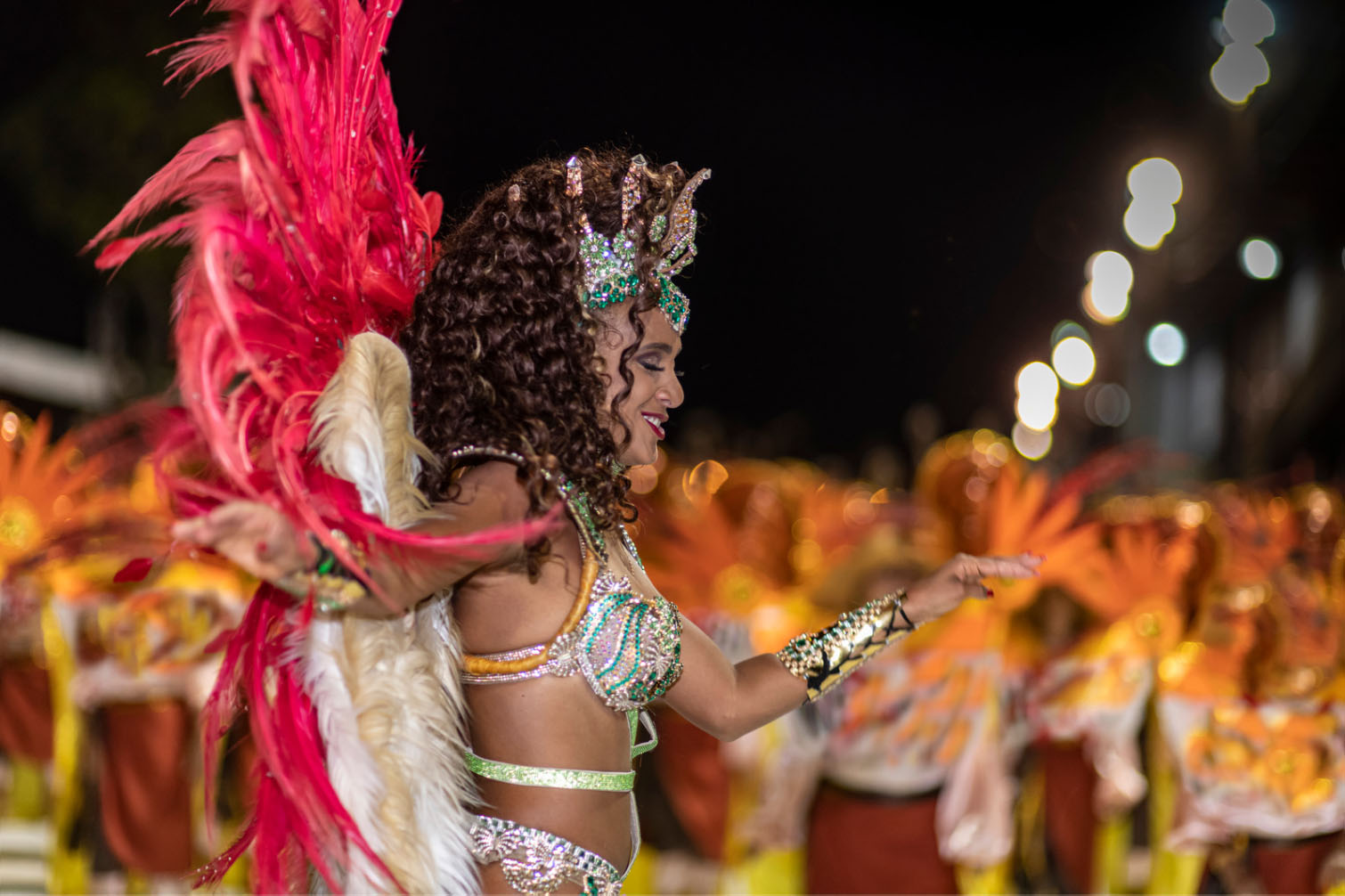 A samba dancer in vibrant costume with large feathered wings performing at the Rio Carnival, with a crowd of dancers in the background.