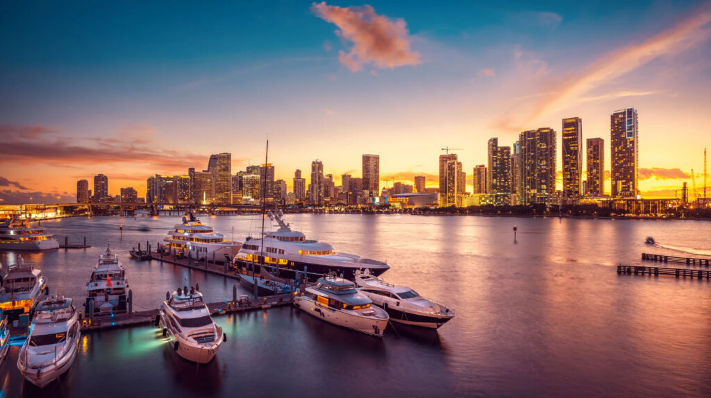 A stunning view of Miami’s skyline at sunset, with luxury yachts docked in the marina and the vibrant city lights reflecting on the water.