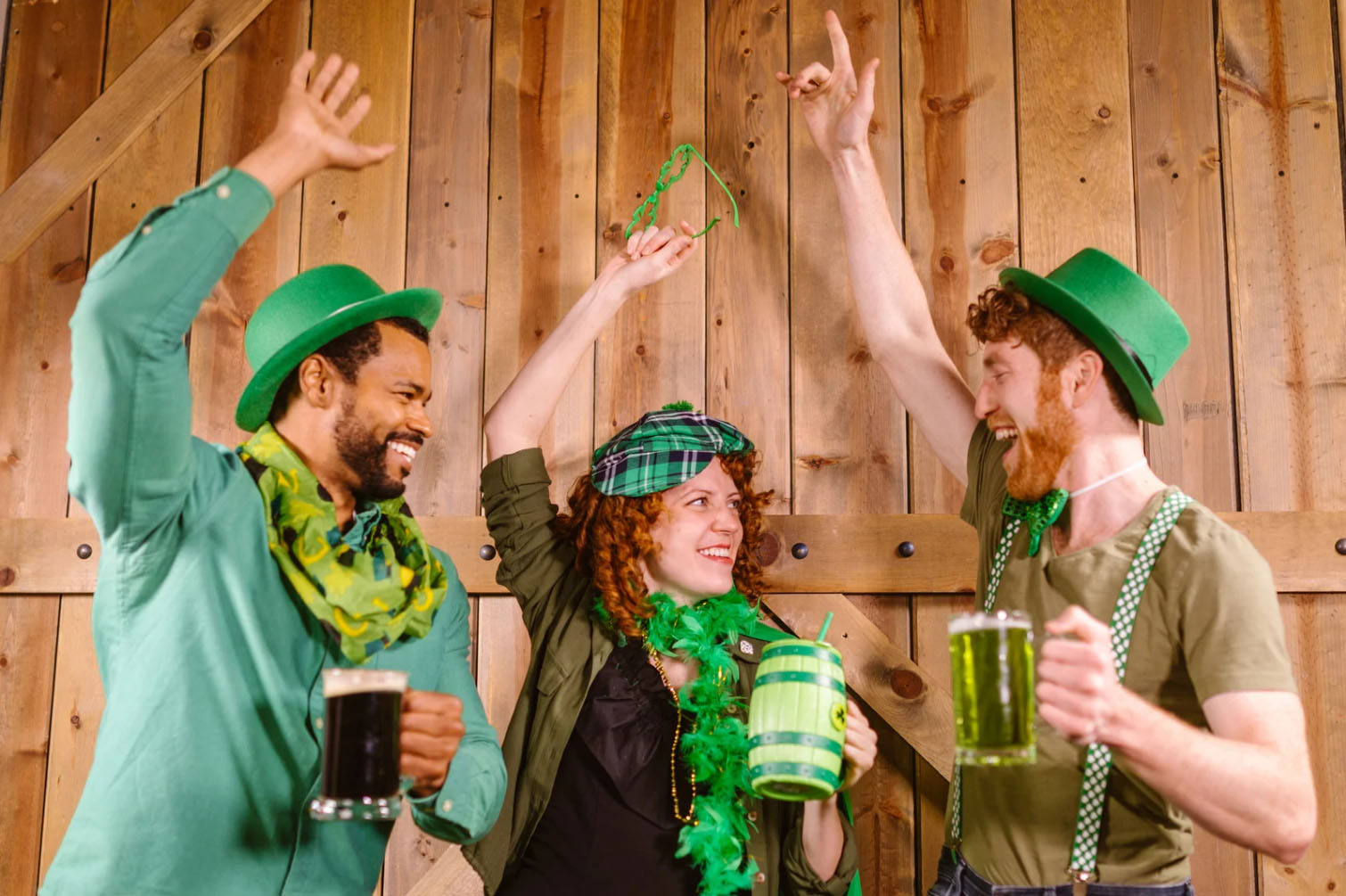Three people celebrating St. Patrick's Day in festive attire, including green hats, scarves, and beads, raising drinks and dancing in front of a wooden background.