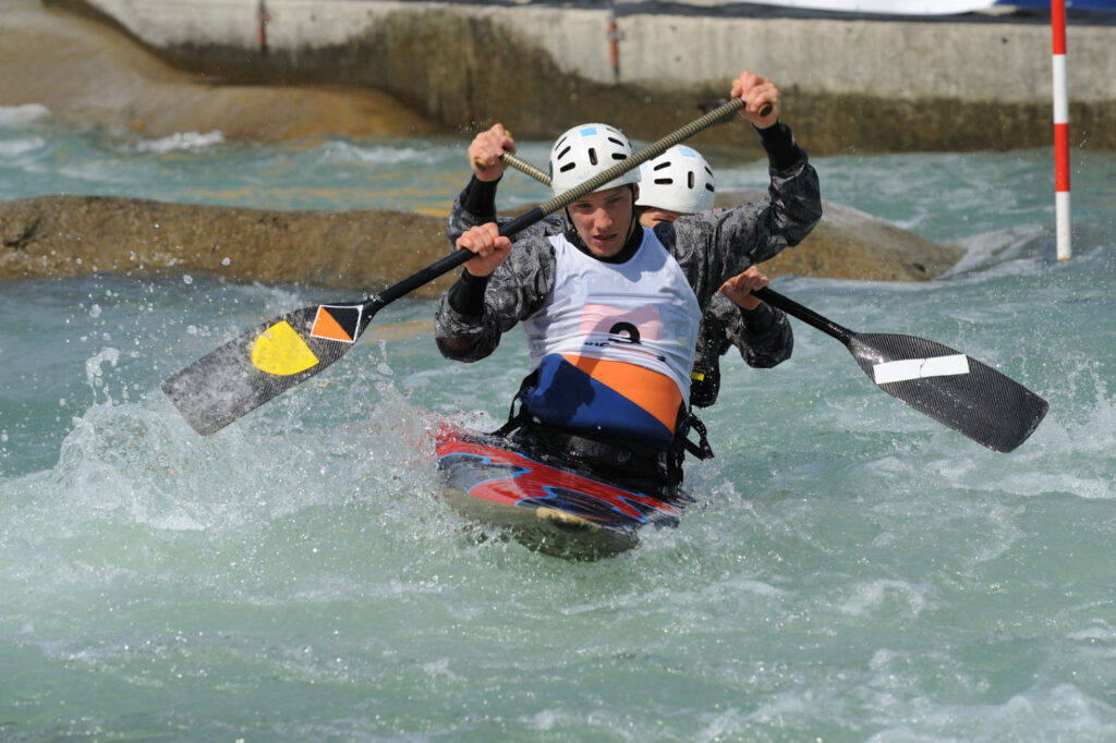 Two athletes navigating a rapid during a white-water kayaking competition, paddling through the course in a canoe with safety gear.