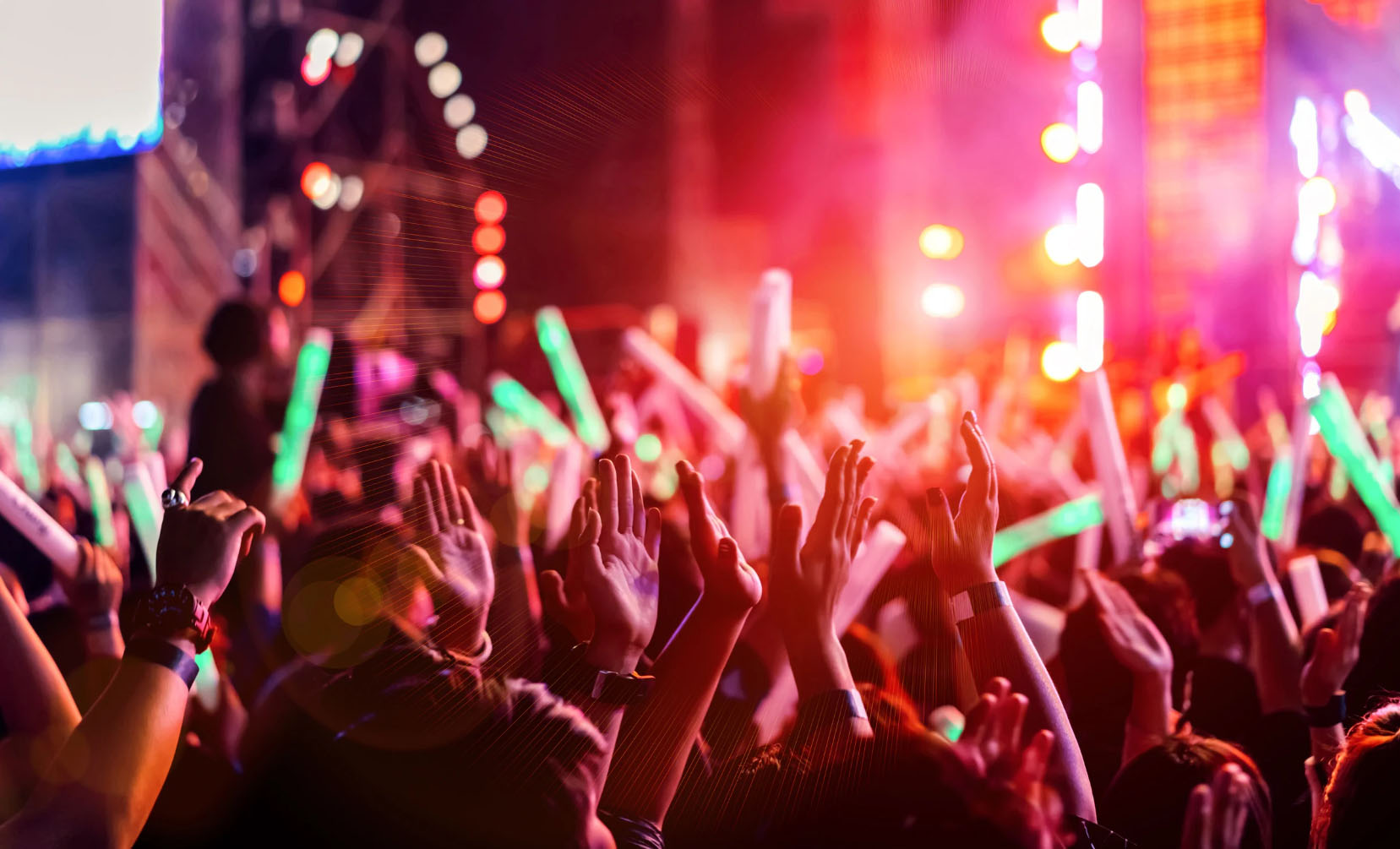 A vibrant crowd at a live concert or music festival with hands raised in the air, glowing light sticks in hand, illuminated by colorful stage lights.