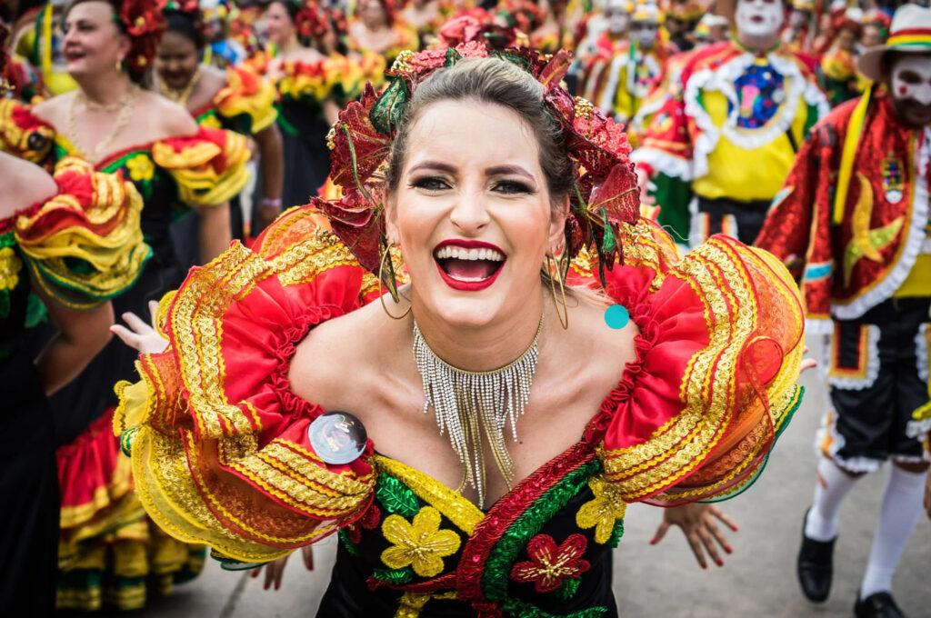 A vibrant participant in the Barranquilla Carnival wearing a colorful traditional dress, smiling widely while dancing in the parade, with other carnival performers in the background.