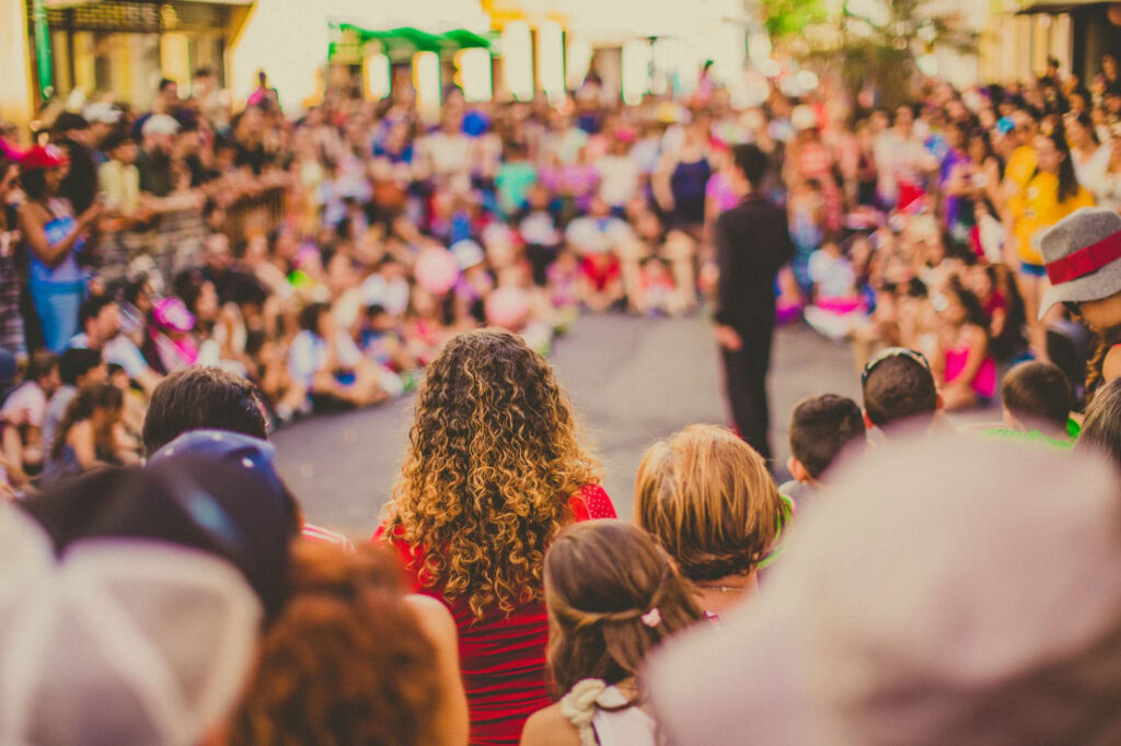 A crowd of people gathered in an outdoor setting watching a performance. The image is taken from behind the audience, focusing on the vibrant crowd and a performer in the distance.