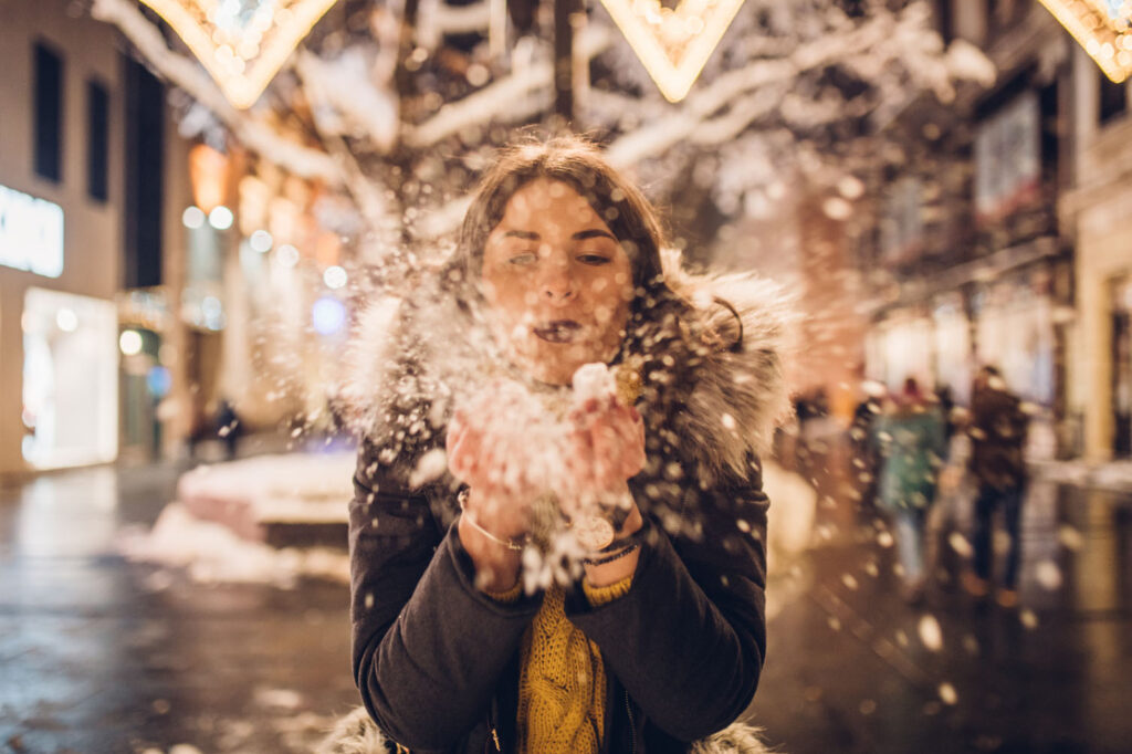 A woman in a winter coat joyfully blowing snowflakes in the air while standing on a snow-covered street, with festive lights and blurred people in the background.