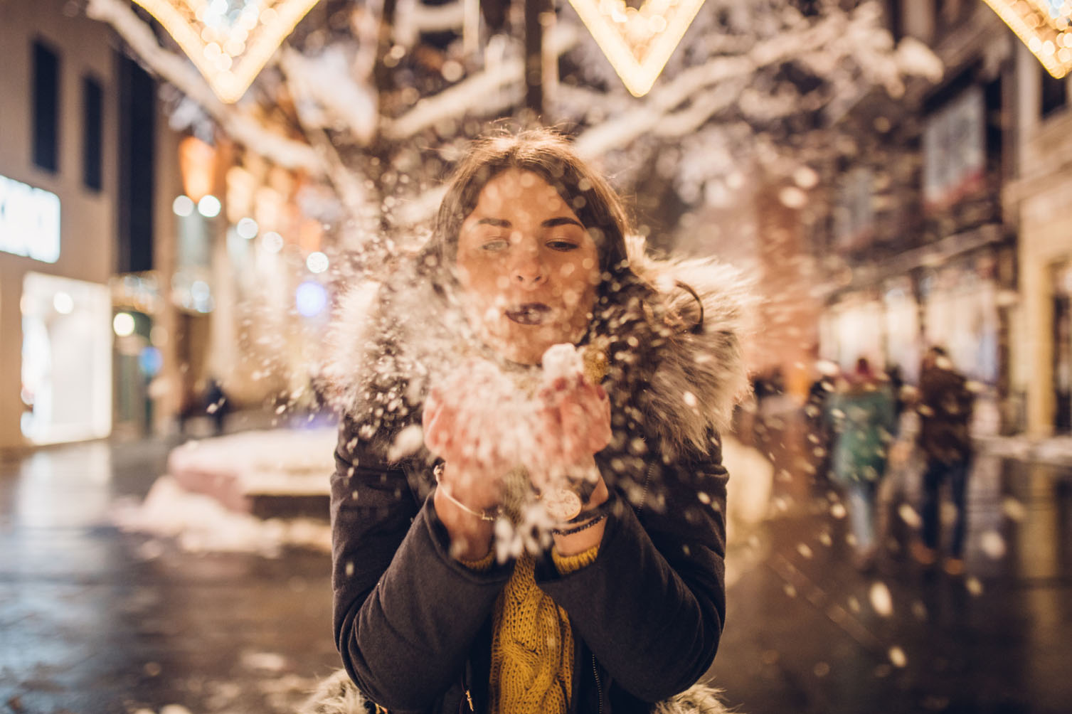 A woman in a winter coat joyfully blowing snowflakes in the air while standing on a snow-covered street, with festive lights and blurred people in the background.
