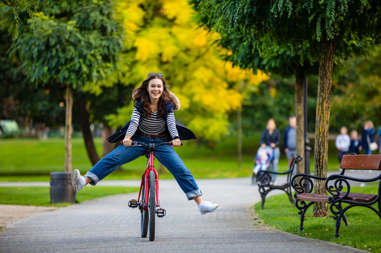 A young woman riding a bicycle in a park, smiling and having fun while balancing on the bike, with a background of trees and people walking.