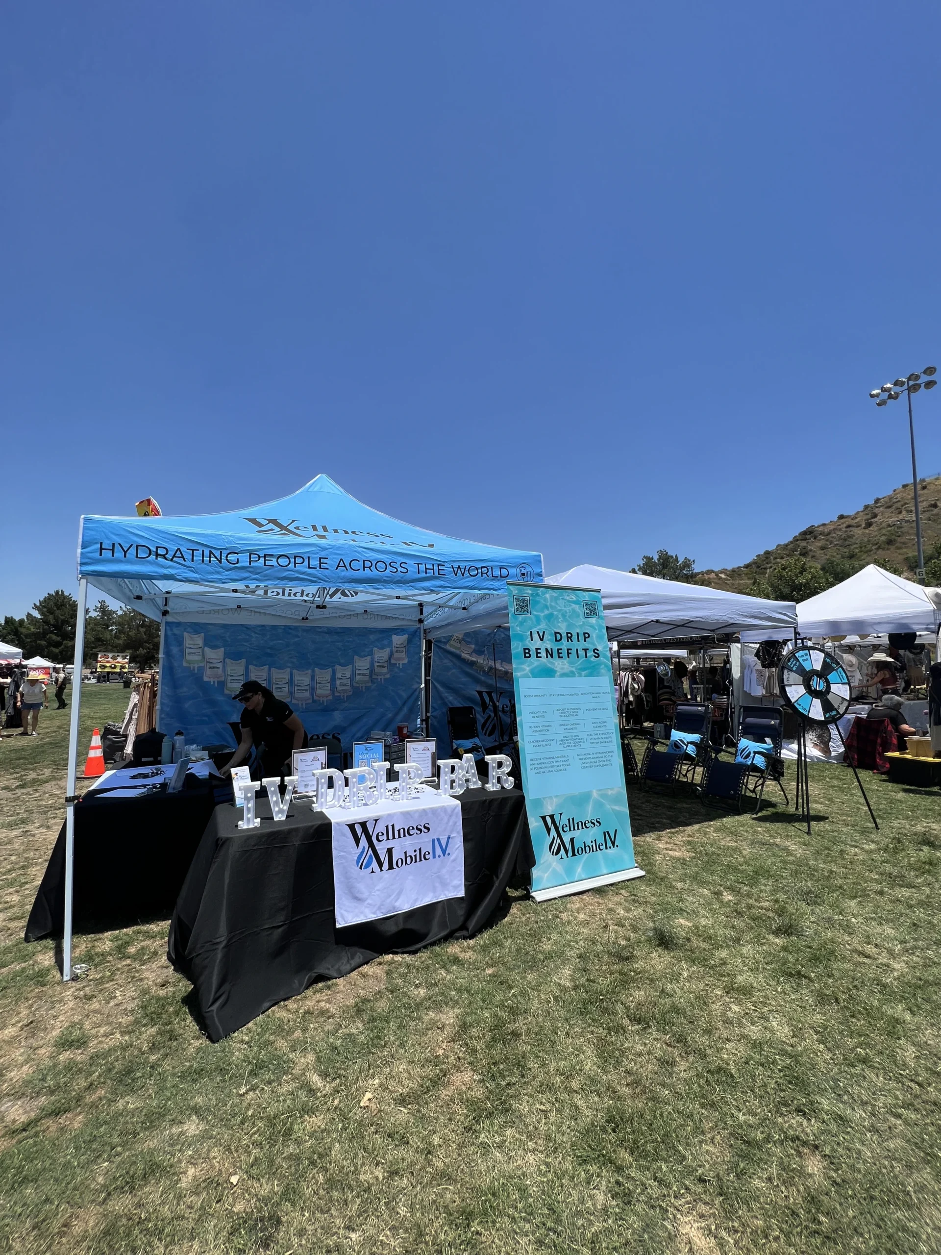 Outdoor event booth for Wellness Mobile IV featuring a blue canopy, 'IV DRIP BAR' marquee sign, and a vertical banner listing benefits on a grassy field.